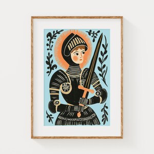 May include: Illustration of a person in black armor holding a sword, set against a light blue background. The artwork features a gold halo and decorative black foliage. The framed print has a wooden frame.