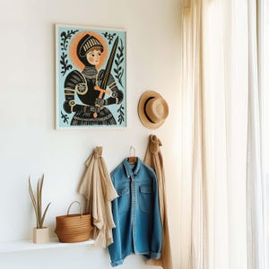 May include: A framed art print of a medieval knight in black armor, holding a sword, set against a light blue background with an orange halo. A straw hat and denim jacket hang on the wall below the artwork, along with a woven basket and a plant.