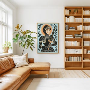 May include: A framed art print featuring a medieval knight in black armor holding a sword against a light blue background. The artwork has a gold frame and is displayed in a living room with a leather sofa and wooden bookshelf.