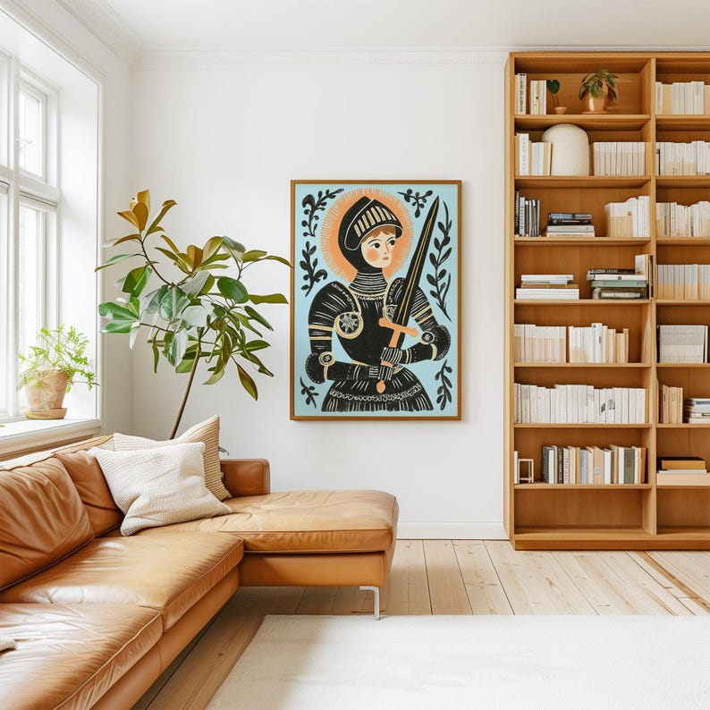 May include: A framed art print featuring a medieval knight in black armor holding a sword against a light blue background. The artwork has a gold frame and is displayed in a living room with a leather sofa and wooden bookshelf.