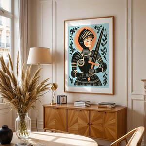May include: A framed art print featuring a stylized illustration of a figure in armor holding a sword. The artwork has a light blue background with black and orange accents. The print is displayed in a room with a wooden cabinet and a large window.