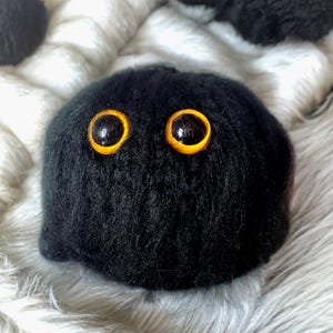 May include: Black fluffy plush toy with large yellow eyes. The toy is shaped like a sphere and has a soft, furry texture.