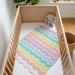K&ouml;nnte beinhalten: Eine Babydecke mit Zickzackmuster in Pastellfarben in einem Holzbettchen. Die Decke hat Reihen in Rosa, Gelb, Gr&uuml;n und Lila. Ein graues Stoffkaninchen sitzt oben im Bettchen. Das Bettchen ist aus hellem Holz.