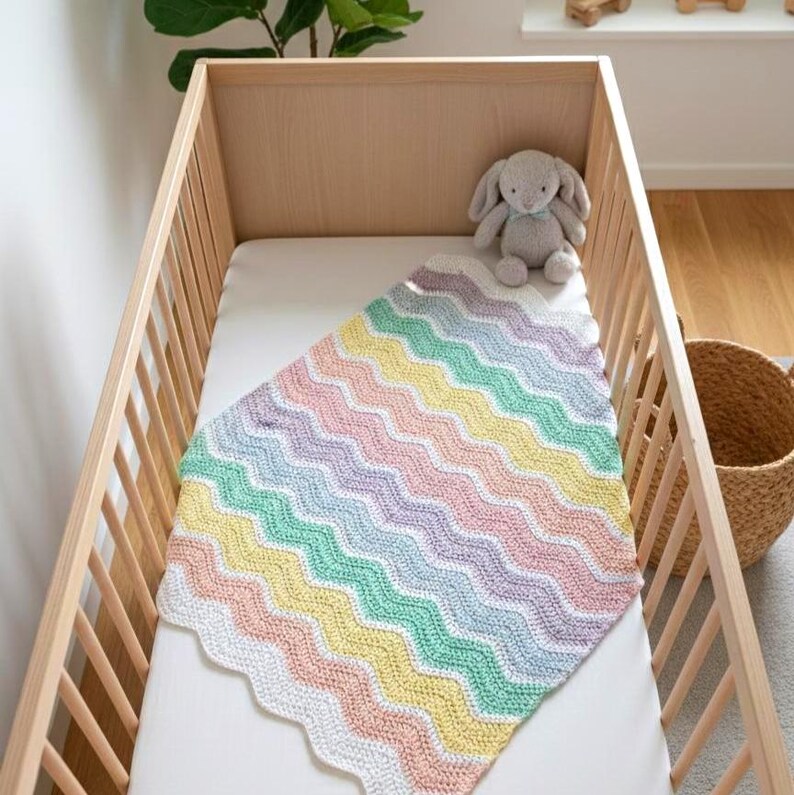 K&ouml;nnte beinhalten: Eine Babydecke mit Zickzackmuster in Pastellfarben in einem Holzbettchen. Die Decke hat Reihen in Rosa, Gelb, Gr&uuml;n und Lila. Ein graues Stoffkaninchen sitzt oben im Bettchen. Das Bettchen ist aus hellem Holz.