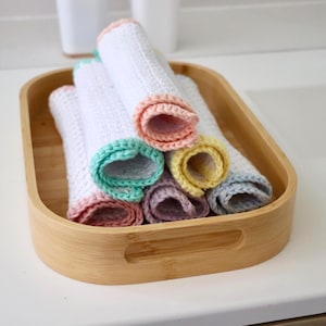 Può includere: Un vassoio di legno contiene diversi asciugamani bianchi arrotolati con bordi all'uncinetto colorati. I bordi sono nelle tonalità di rosa, verde, giallo, viola e blu. Il vassoio è in legno chiaro.