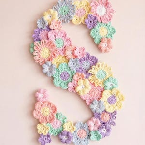 Puede incluir: Una letra "S" de ganchillo en colores pastel, compuesta por varias flores de ganchillo en tonos rosa, amarillo, verde y morado. Las flores est&aacute;n dispuestas para formar la letra sobre un fondo rosa claro. El dise&ntilde;o floral hecho a mano es adecuado para la decoraci&oacute;n de la guarder&iacute;a.