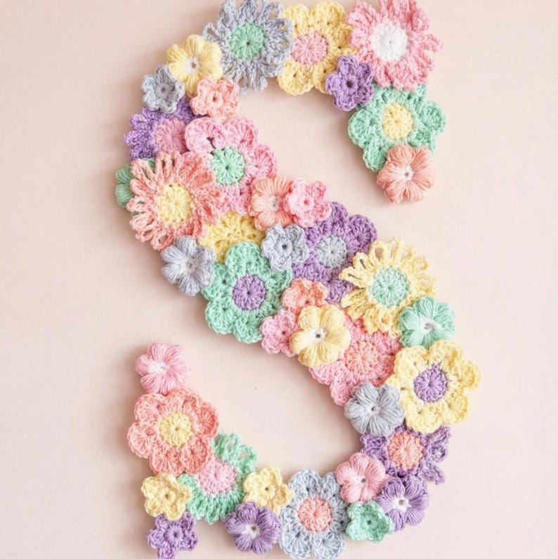 Puede incluir: Una letra "S" de ganchillo en colores pastel, compuesta por varias flores de ganchillo en tonos rosa, amarillo, verde y morado. Las flores est&aacute;n dispuestas para formar la letra sobre un fondo rosa claro. El dise&ntilde;o floral hecho a mano es adecuado para la decoraci&oacute;n de la guarder&iacute;a.