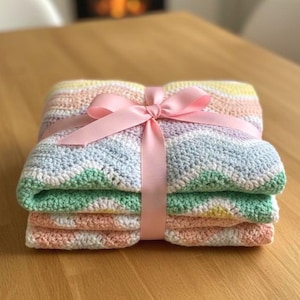 K&ouml;nnte beinhalten: Eine gefaltete, pastellfarbene H&auml;keldecke, die mit einem rosa Band zusammengebunden ist. Die Decke hat Streifen in Wei&szlig;, Gr&uuml;n, Gelb, Rosa und Blau. Die Decke ist ordentlich auf einer Holzoberfl&auml;che gestapelt.