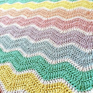 K&ouml;nnte beinhalten: Eine handgefertigte, farbenfrohe H&auml;keldecke mit Zickzackmuster. Die Decke zeigt Reihen in verschiedenen Farben, darunter Gelb, Gr&uuml;n, Rosa und Lila. Die Maschen sind sichtbar und erzeugen eine strukturierte Oberfl&auml;che. Das Gesamtdesign ist eine Mischung aus Pastell- und leuchtenden Farben.