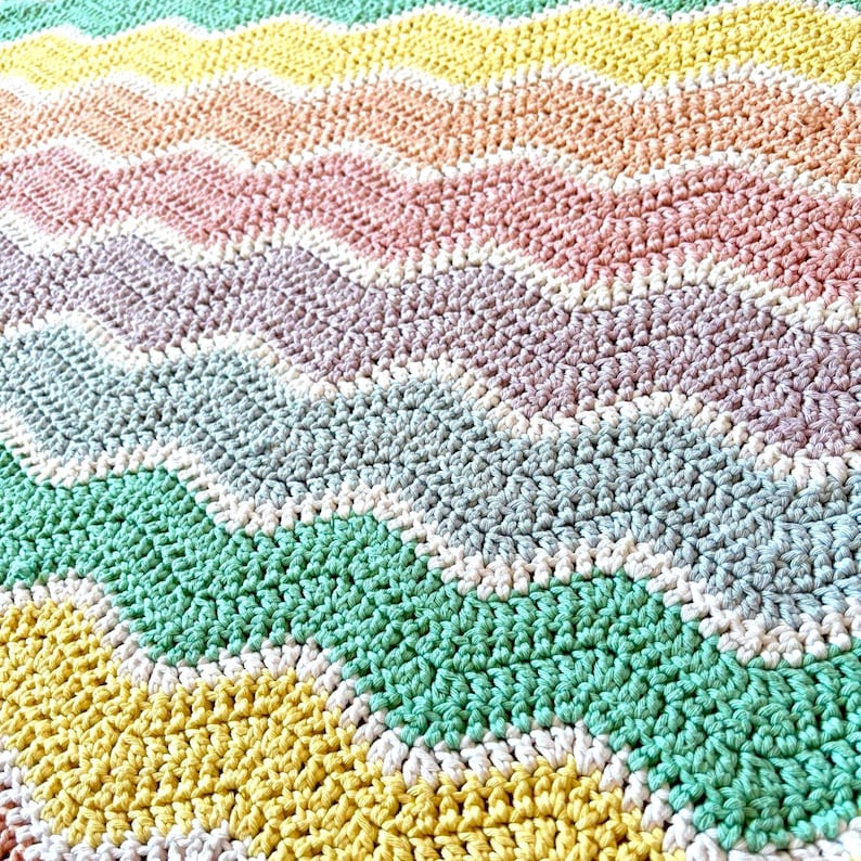 K&ouml;nnte beinhalten: Eine handgefertigte, farbenfrohe H&auml;keldecke mit Zickzackmuster. Die Decke zeigt Reihen in verschiedenen Farben, darunter Gelb, Gr&uuml;n, Rosa und Lila. Die Maschen sind sichtbar und erzeugen eine strukturierte Oberfl&auml;che. Das Gesamtdesign ist eine Mischung aus Pastell- und leuchtenden Farben.