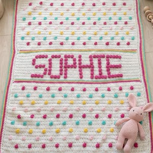 以下が含まれることがあります： カラフルな水玉模様とピンク色の「SOPHIE」という名前が入った、手作りの白いかぎ針編みのベビーブランケット。ブランケットはピンク、黄色、ティール色の縁取りがあり、お揃いのピンク色のぬいぐるみウサギが付いています。パーソナライズされた居心地の良いベビーギフト。