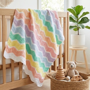 K&ouml;nnte beinhalten: Eine pastellfarbene, geh&auml;kelte Babydecke mit Wellenmuster liegt &uuml;ber einem Holzbettchen. Die Decke hat Streifen in Pfirsich, Gelb, Gr&uuml;n, Blau und Lavendel mit einem wei&szlig;en, gewellten Rand. Unter dem Bettchen steht ein Korb mit Spielzeug.