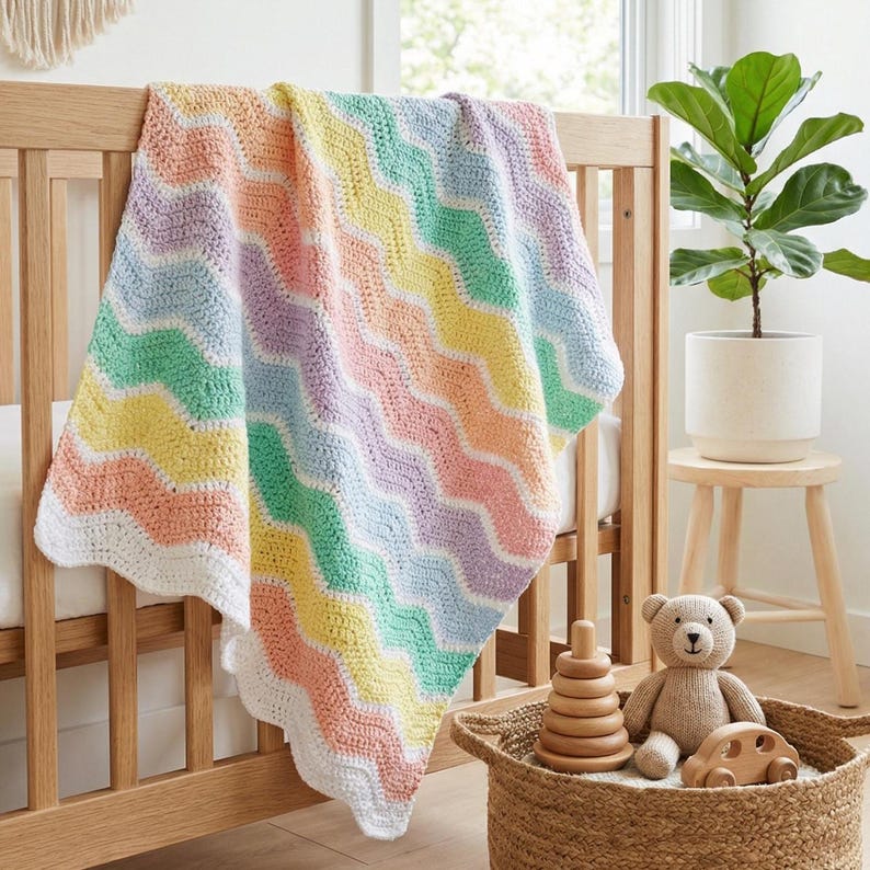 K&ouml;nnte beinhalten: Eine pastellfarbene, geh&auml;kelte Babydecke mit Wellenmuster liegt &uuml;ber einem Holzbettchen. Die Decke hat Streifen in Pfirsich, Gelb, Gr&uuml;n, Blau und Lavendel mit einem wei&szlig;en, gewellten Rand. Unter dem Bettchen steht ein Korb mit Spielzeug.