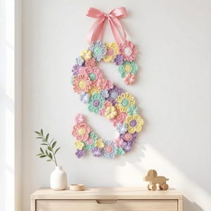 Puede incluir: Una letra "S" de ganchillo en colores pastel, adornada con flores de ganchillo rosas, amarillas, moradas y verdes. Cuelga de una pared con una cinta rosa. Un peque&ntilde;o perro de madera y un jarr&oacute;n est&aacute;n sobre una superficie de madera.