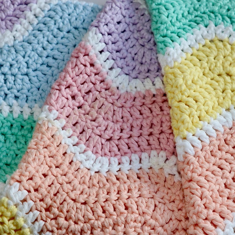 K&ouml;nnte beinhalten: Nahaufnahme einer handgefertigten H&auml;keldecke in Pastellfarben. Die Decke hat ein Streifenmuster mit Reihen in Wei&szlig;, Rosa, Pfirsich, Gelb, Gr&uuml;n und Blau. Die Textur der H&auml;kelstiche ist sichtbar.