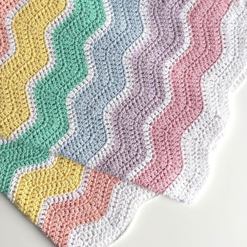 K&ouml;nnte beinhalten: Eine handgefertigte H&auml;keldecke mit Wellenmuster in Pastellfarben. Die Decke hat Streifen in Gelb, Gr&uuml;n, Hellblau, Lavendel, Rosa und Wei&szlig;. Die Kanten sind mit Muschelmuster versehen, was der weichen, strukturierten Decke eine dekorative Note verleiht.