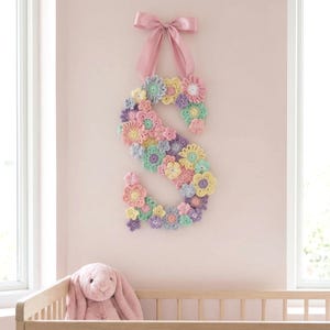 Puede incluir: Una letra "S" decorativa de ganchillo, adornada con flores en tonos pastel, suspendida por un lazo de cinta rosa. La pared est&aacute; pintada de un rosa suave. Un conejito de peluche es visible en primer plano, sugiriendo una habitaci&oacute;n infantil.