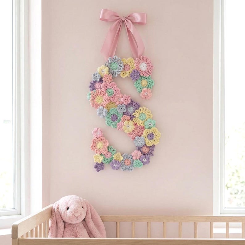 Puede incluir: Una letra "S" decorativa de ganchillo, adornada con flores en tonos pastel, suspendida por un lazo de cinta rosa. La pared est&aacute; pintada de un rosa suave. Un conejito de peluche es visible en primer plano, sugiriendo una habitaci&oacute;n infantil.