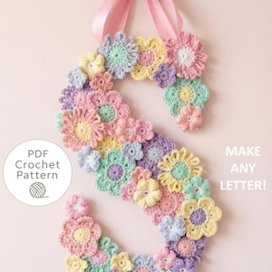 Può includere: Una lettera "S" all'uncinetto adornata con fiori dai colori pastello, sospesa da un nastro rosa. Il design presenta una varietà di forme di fiori nelle tonalità del rosa, giallo, blu e viola. Il testo a sinistra recita "PDF Crochet Pattern" e a destra "MAKE ANY LETTER!"