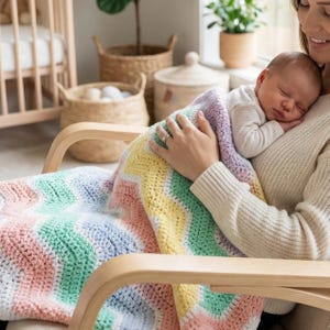 K&ouml;nnte beinhalten: Eine Babydecke aus H&auml;kelarbeit mit Zickzackmuster in Pastellt&ouml;nen von Rosa, Gelb, Gr&uuml;n und Blau. Die Decke ist &uuml;ber einen Holzstuhl drapiert. Ein schlafendes Baby wird von einer Person gehalten, die einen cremefarbenen Pullover tr&auml;gt.