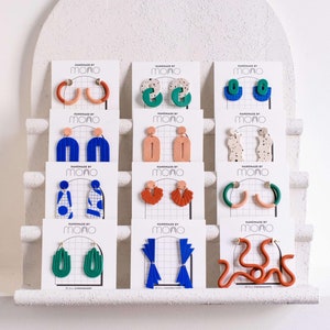 Pode incluir: Uma exibição de brincos de argila polimérica coloridos em cartões brancos. Os brincos são de várias formas e designs, incluindo formas geométricas, abstratas e orgânicas. Os cartões são etiquetados com "Handmade by Mono".