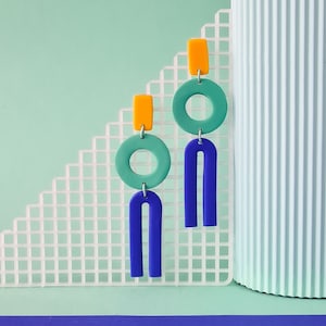 Può includere: Un paio di orecchini geometrici con uno schema di colori blu, verde e arancione. Gli orecchini presentano un cerchio grande, un cerchio più piccolo e una forma curva.