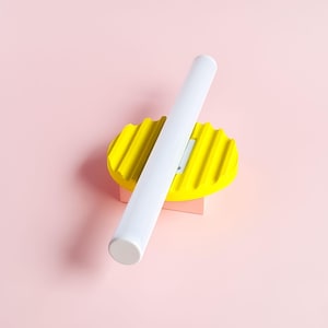 May include: A white cylindrical object rests diagonally across a yellow, ridged oval shape, which is placed on a small pink rectangular block. The background is a solid light pink. The objects are brightly coloured and have a minimalist design.