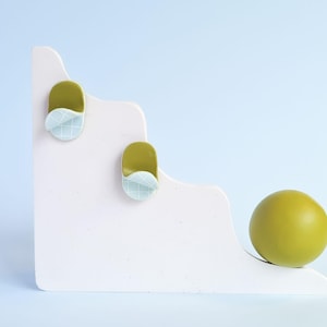 May include: A white, stepped platform with two olive-green and blue decorative elements and a matching sphere. The elements have a curved, grid-patterned design. The sphere is resting on the platform, against a light blue backdrop.
