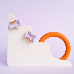 May include: An abstract sculpture with a white stepped base, an orange ring, and two purple shapes with a grid pattern. The background is a soft purple. The sculpture is a modern art piece.