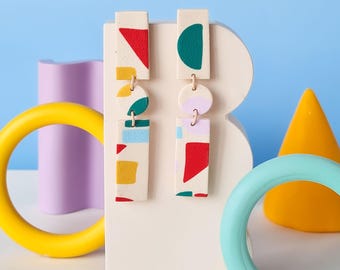 May include: A pair of geometric patterned earrings with white, red, green, yellow, and blue colors. The earrings are made of polymer clay and have a rectangular shape.