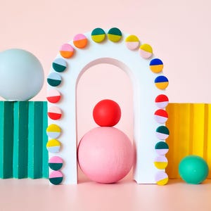 May include: An abstract arrangement of geometric shapes in a pastel palette. A white arch is decorated with two-tone circular pieces. Spheres in blue, pink, red, and green are placed around the scene against a pink background, creating a playful aesthetic.
