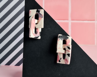 May include: Two abstract polymer clay earrings. Each earring features a rectangular shape with a cutout design. The earrings have a speckled pattern in shades of pink, black, and white. The earrings are displayed on a black triangle against a pink and white background.
