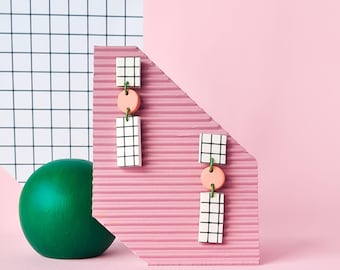 May include: A pair of geometric earrings with black and white checkered squares, pink circles, and green accents. The earrings are displayed on a pink background.