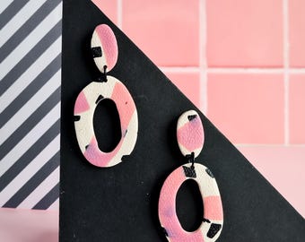 May include: A pair of abstract dangle earrings. The earrings feature an oval shape with a smaller oval at the top. The earrings are white with pink and black abstract patterns. The background includes a black triangle, pink tiles, and a black and white striped pattern.