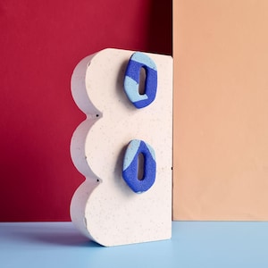 Op de afbeelding: Twee abstracte blauwe en lichtblauwe oorbellen met een textuuroppervlak worden getoond op een witte, golvende, getextureerde blok. De achtergrond heeft een rode muur, een perzikkleurige muur en een blauw oppervlak.