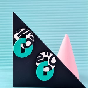 Puede incluir: Dos pendientes con estampado en blanco y negro con un centro verde turquesa sobre una superficie triangular negra sobre un fondo azul claro.