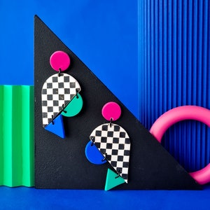 Puede incluir: Un par de pendientes con estampado de tablero de ajedrez con formas geométricas rosas, azules y verdes. Los pendientes están sobre un fondo negro.