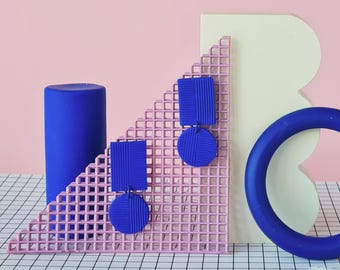 May include: A pair of cobalt blue dangle earrings, each featuring a rectangular top piece connected to a circular bottom piece. Both components have a fine, vertical ribbed texture. The earrings are displayed against a light pink background.