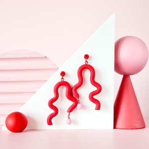 Puede incluir: Pendientes abstractos de color rojo brillante, con un diseño ondulado y pequeñas cuentas rosas. Los pendientes se exhiben contra una forma geométrica blanca y un fondo rosa, con una esfera roja y una esfera rosa sobre un cono.