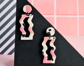May include: A pair of abstract earrings with a pink, white, and black pattern. The earrings have a circular top and a wavy, rectangular bottom. They are displayed on a black triangle against a pink tiled background.