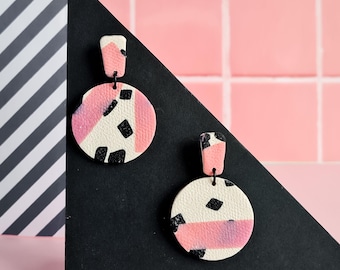 May include: A pair of abstract geometric earrings. Each earring features a rectangular top and a circular pendant. The pendants are white with pink and black abstract shapes. The earrings are displayed on a black triangle.