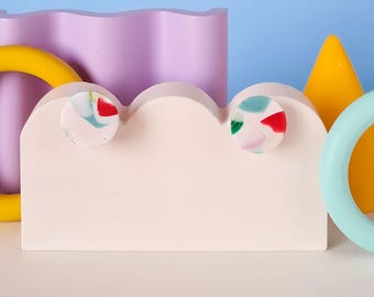May include: A light pink, cloud-shaped display stand with two circular, colorful earrings. The earrings have red, green, and blue accents. Other geometric shapes in yellow, purple, and light blue are in the background.