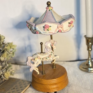 May include: A carousel music box featuring a white horse with a floral garland, standing on a round wooden base. The canopy is decorated with pastel pink and yellow roses, blue ribbons, and a lavender roof, supported by gold-colored metal poles.