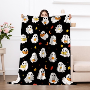 May include: A black Halloween-themed blanket featuring a repeating pattern of white cartoon ghosts holding pumpkins and books. The design includes yellow stars, orange leaves, and crescent moons. The blanket is held up by a person.