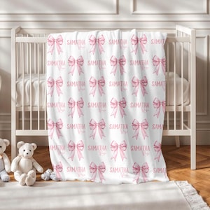 Könnte beinhalten: Eine weiße Babydecke mit einem sich wiederholenden Muster aus rosa Schleifen und dem Namen "SAMATHA Rose" in zarter Schrift. Die Decke ist über ein weißes Kinderbett drapiert, mit Stofftieren sichtbar.