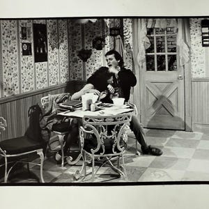 May include: Black and white photograph of a vintage interior scene. A couple is seated at a small, ornate table. The room features patterned wallpaper, a checkered floor, and a decorative chair.