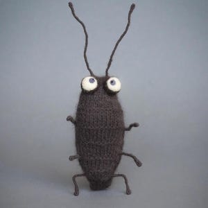 May include: A dark brown knitted insect figurine with a fuzzy texture, featuring two long antennae, four thin legs, and two large, round white eyes with dark pupils. The body has a ribbed, yarn-like appearance and stands upright on its own.