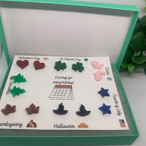 May include: A white box with a green lid containing a set of 10 pairs of earrings. Each pair is decorated with a different holiday theme, including hearts for Valentine's Day, shamrocks for St. Patrick's Day, bunnies for Easter, Christmas trees for Christmas, turkeys for Thanksgiving, witches' hats for Halloween, and stars for the Fourth of July. The box has a calendar image with the text "Earrings for every holiday!" printed on it.