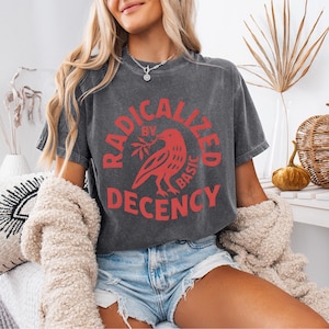 May include: Dark gray t-shirt with the text "RADICALIZED BY BASIC DECENCY" in red. The design includes a red bird holding a sprig. The shirt is a casual wear item.