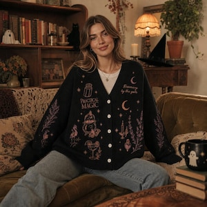 May include: A black button-down cardigan featuring pink graphic prints of a cauldron, a cocktail glass, a candle, a moth, and the text "Practical Magic" and "Rosemary by the gate" along with various botanical and celestial motifs.